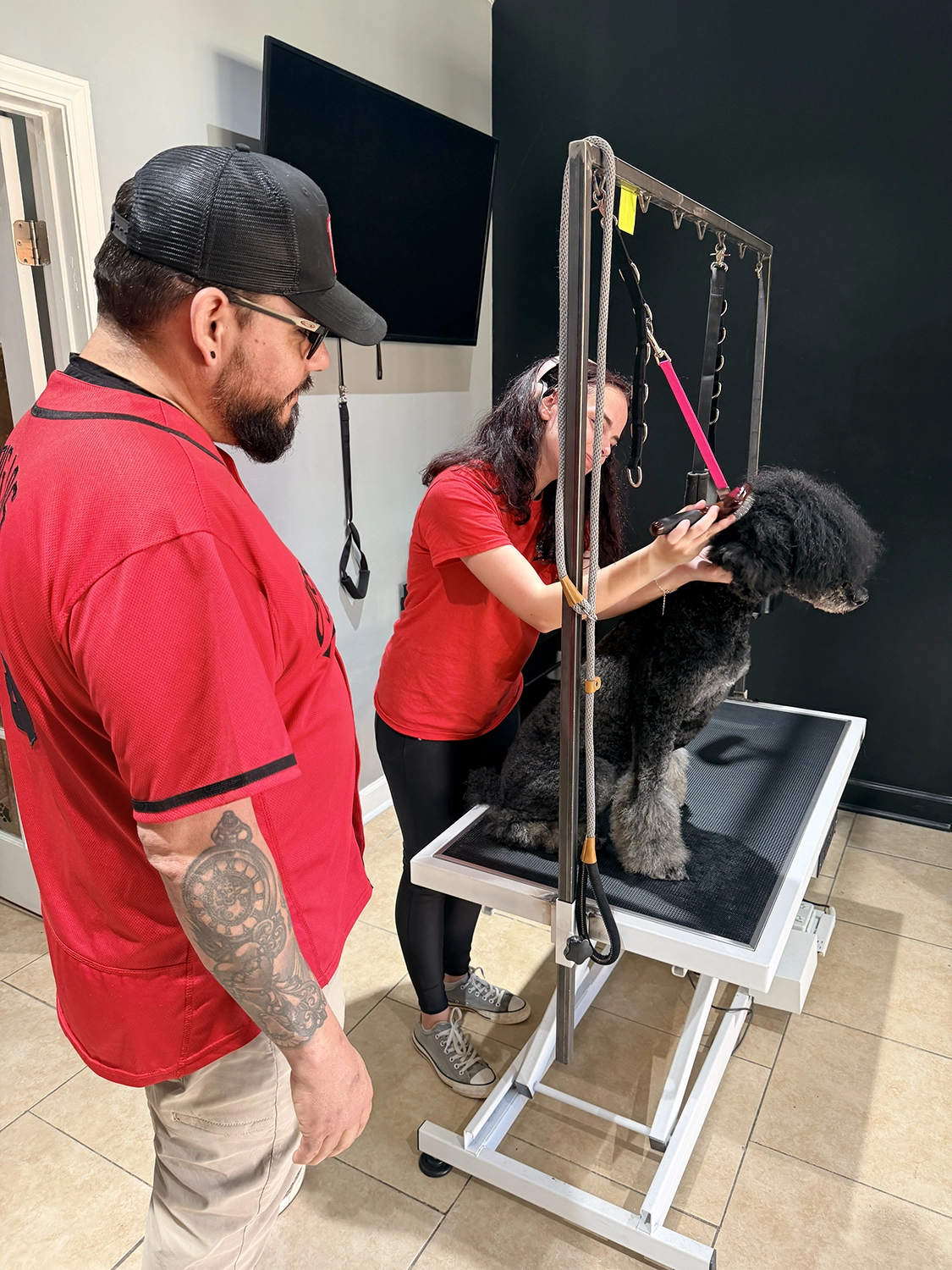 Dog Grooming 2 Dog grooming session at Off The Chain K9 Academy, featuring a groomer in red shirt using a grooming table with a black dog, while a client observes, emphasizing a calm and structured environment.