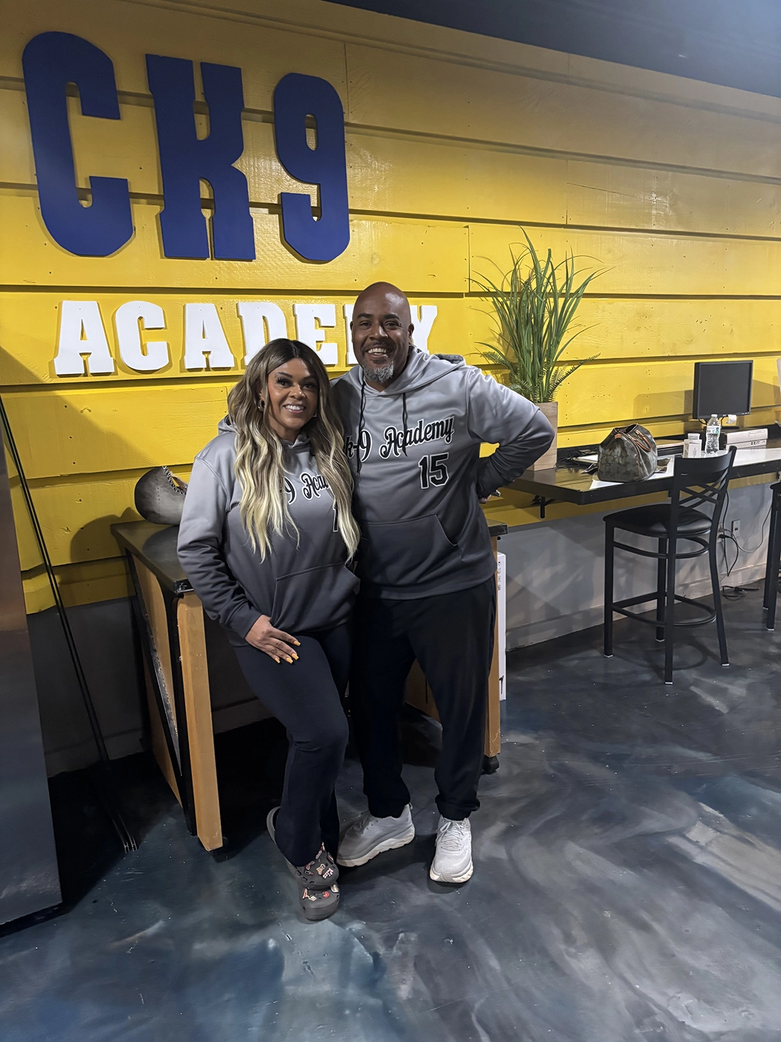 About 3 Darrell and Kim, founders of Off The Chain K9 Academy, smiling in front of the academy's colorful wall, showcasing their commitment to dog training and education.