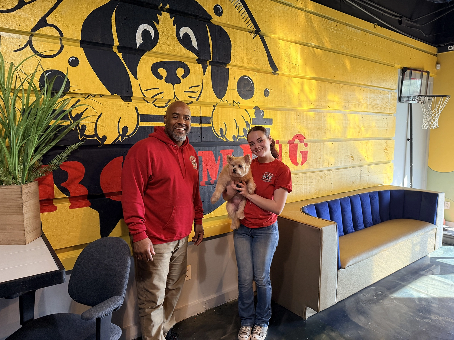 Professional dog grooming instructors and student with a dog in a grooming academy setting, featuring a vibrant yellow wall with a dog illustration, emphasizing hands-on training in dog grooming certification.