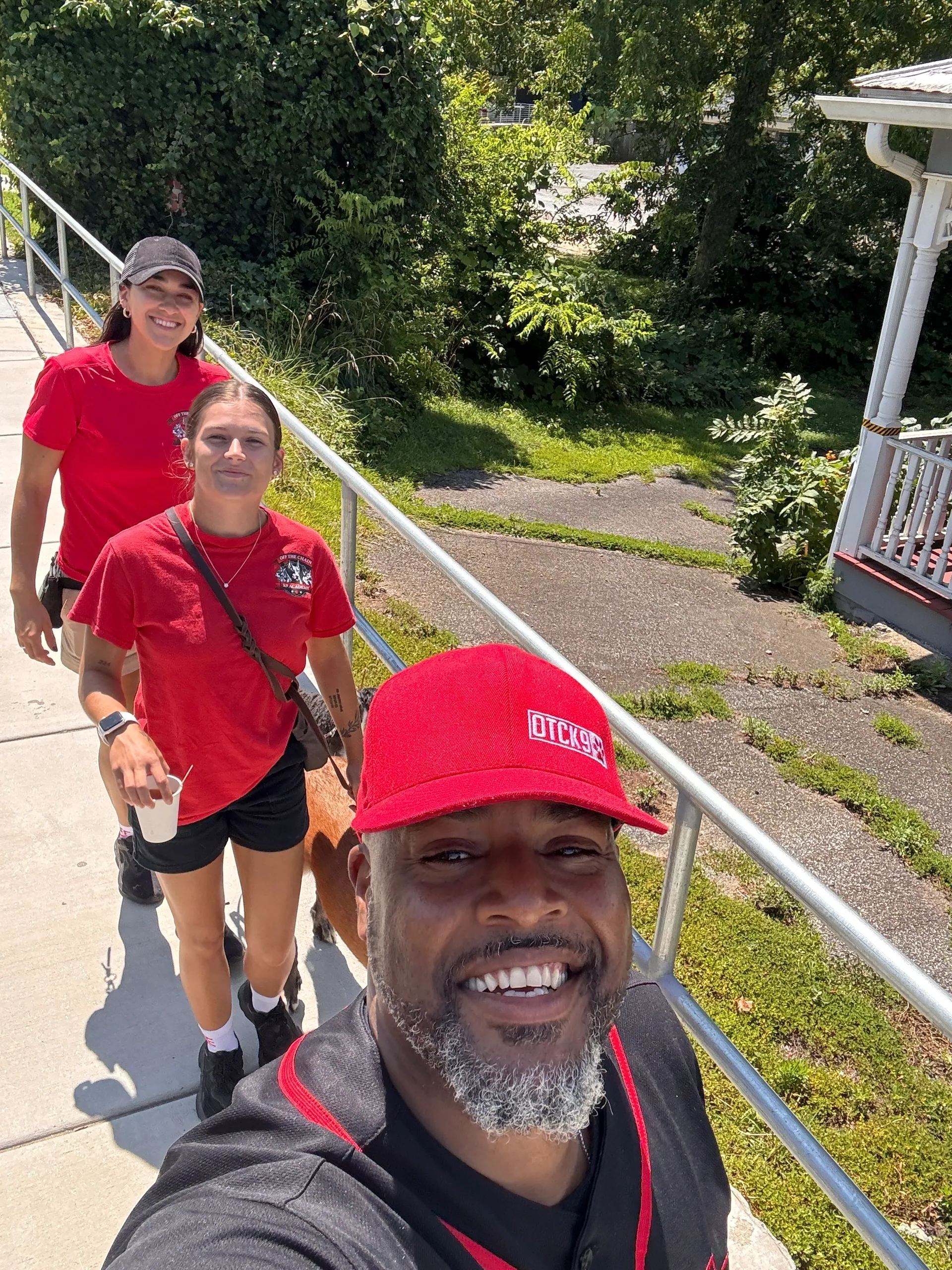 Two dog trainers in red OTCK9 Academy shirts walking along a path with a dog, showcasing a friendly training environment and a focus on real-world dog training.