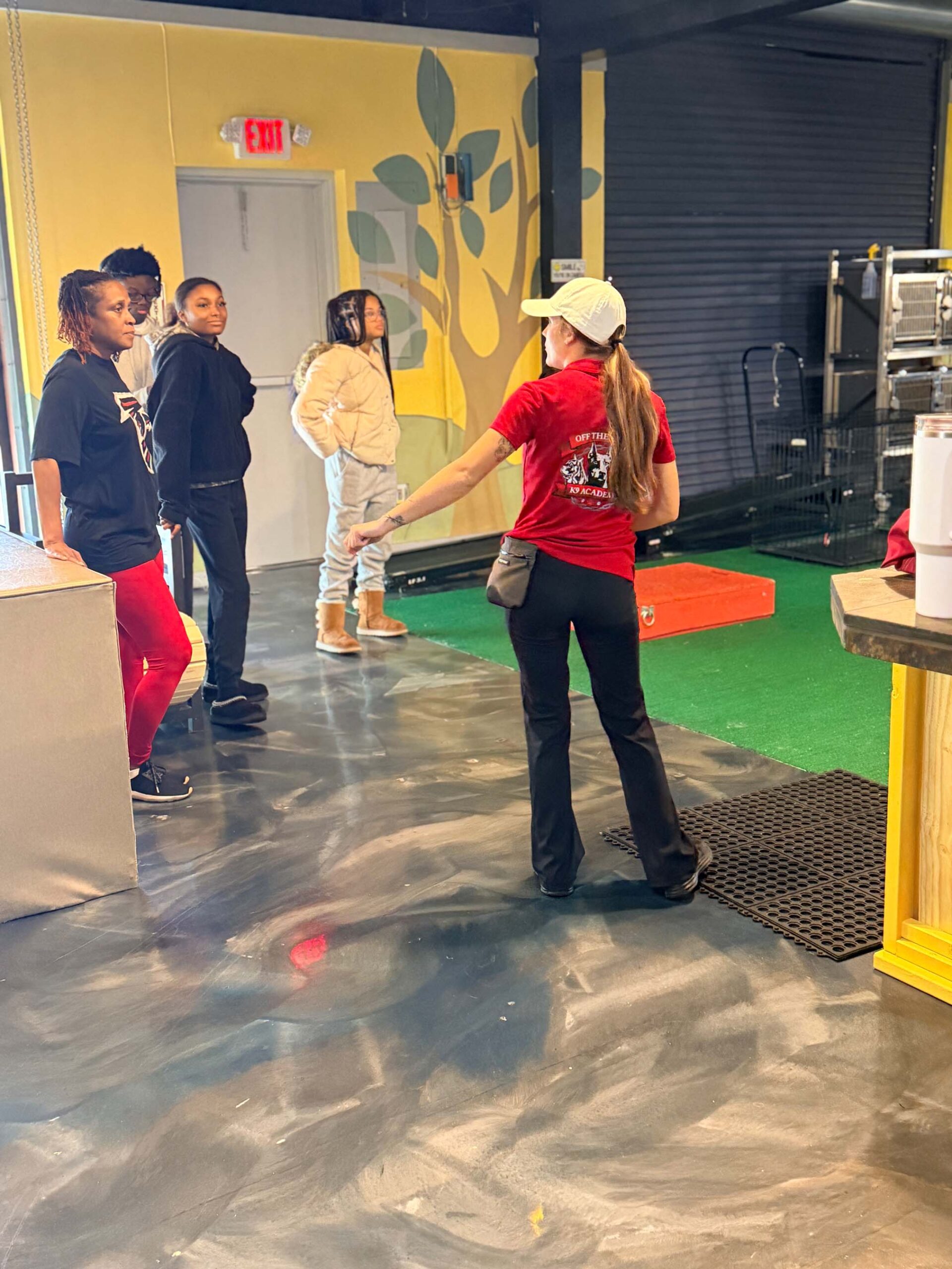Dog Board and Train Program 6 A woman in a red shirt speaks to four people standing near a counter in an indoor space with a painted floor and a yellow wall.