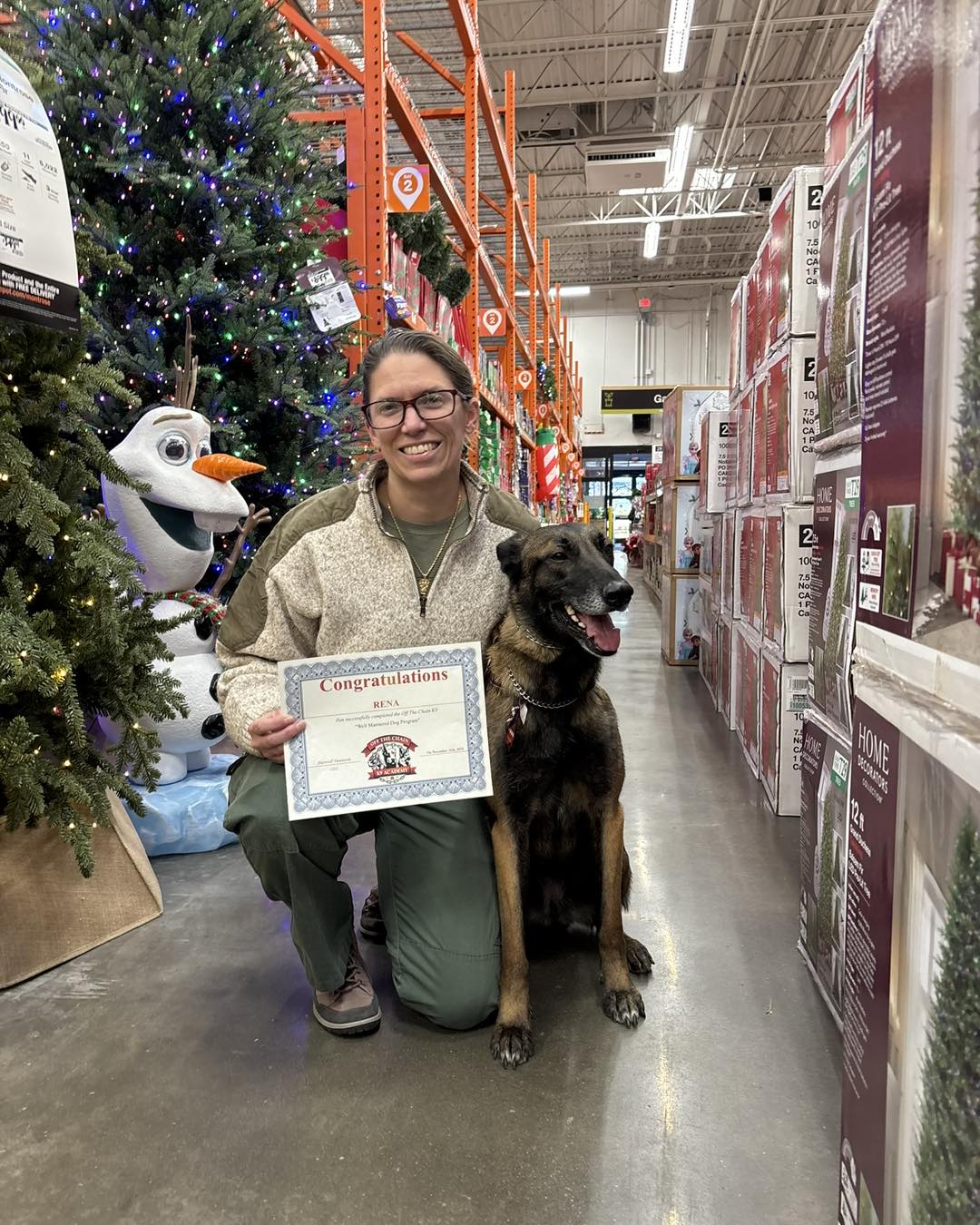 A person kneels next to a dog in a store aisle, holding a certificate after completing a dog training certification in Woodstock GA, with artificial Christmas trees and holiday decorations displayed around them.