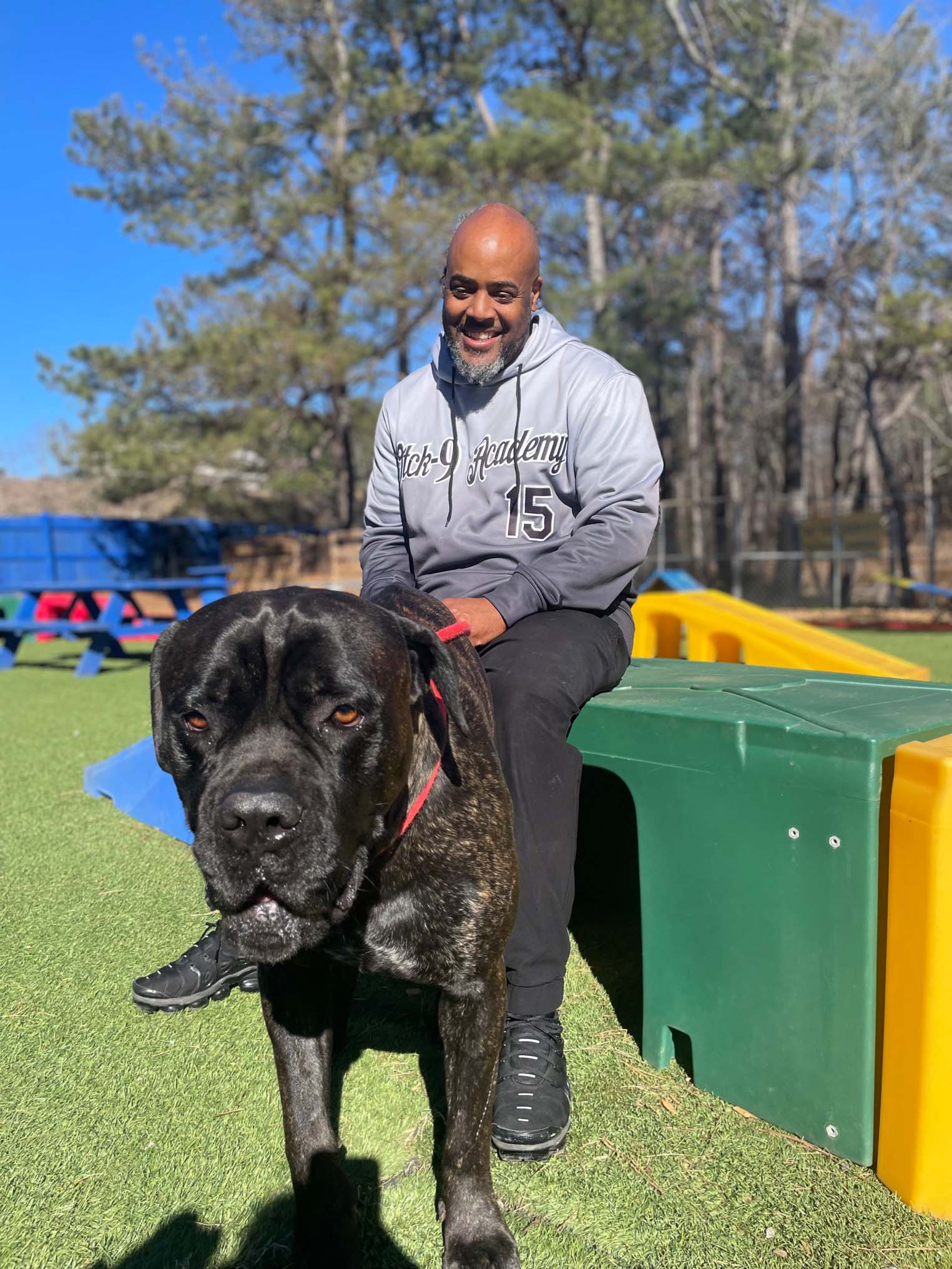Doggy Day School 5 Man smiling while sitting on a colorful play structure with a large dog in a training environment, emphasizing structured dog training and socialization at Off The Chain K9 Academy.