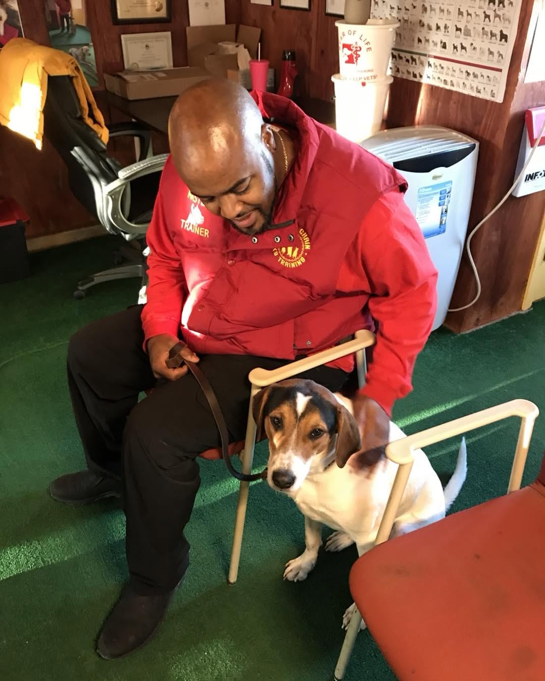 A man in a red jacket sits indoors on a chair, holding a leash attached to a brown and white dog beside him, possibly practicing behavior modification techniques.