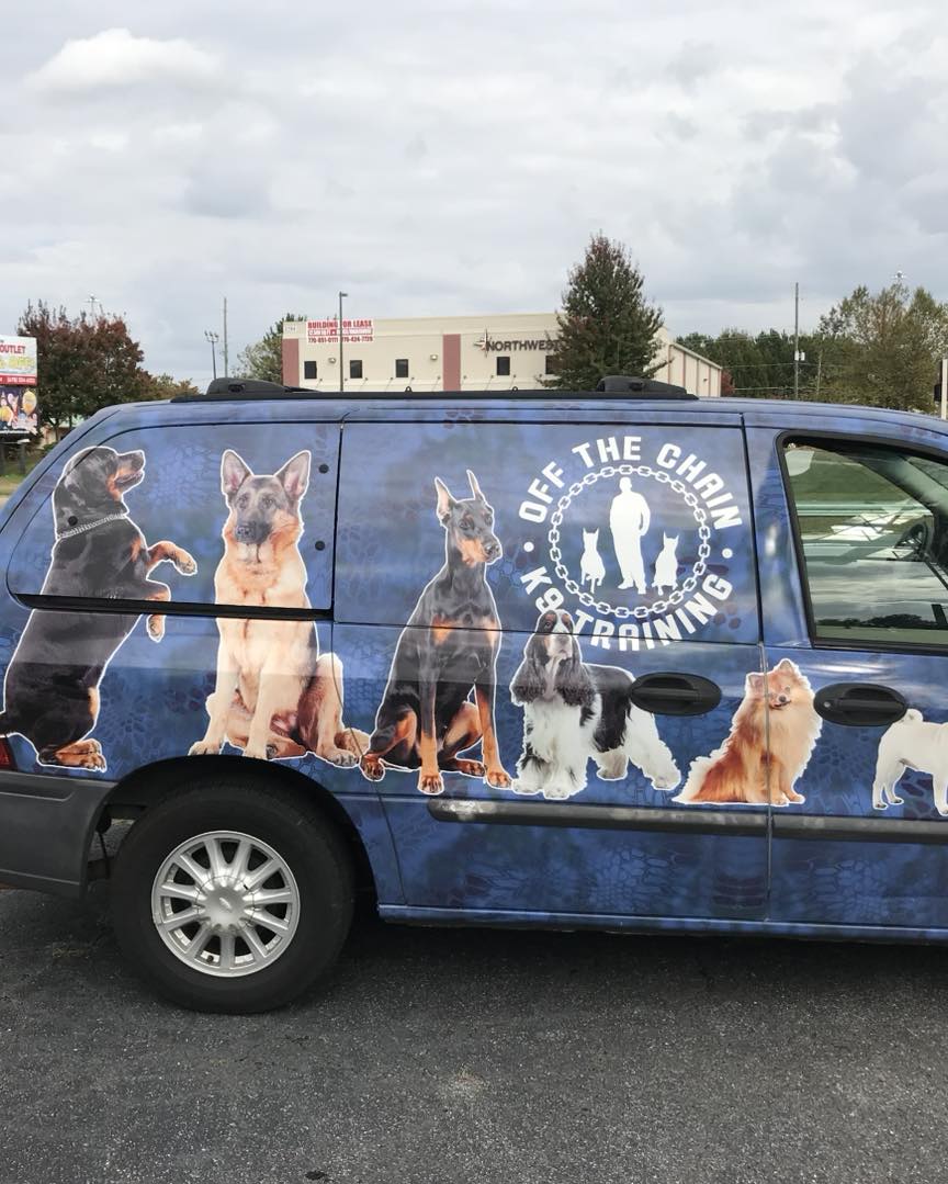 A blue van with the "Off The Chain K9 Training" logo and images of various dog breeds, specializing in behavior modification, is parked in a lot.