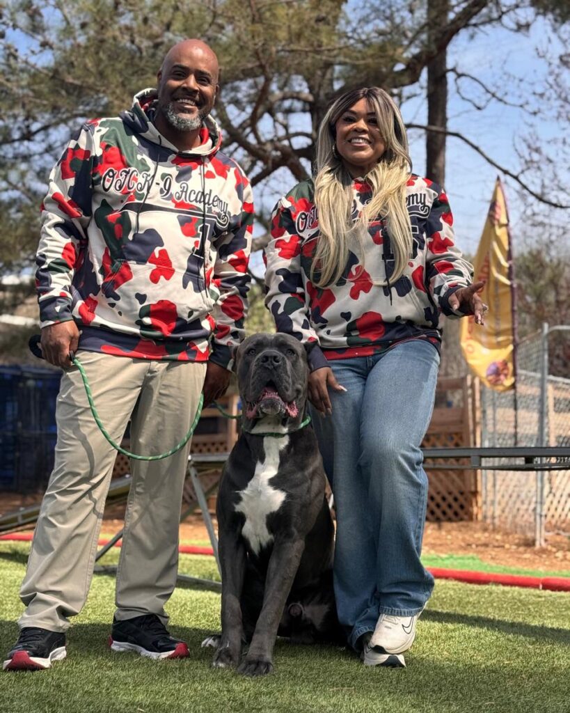 Couple in matching OTCK9 Academy hoodies with a large dog sitting on grass, showcasing a friendly dog training environment.