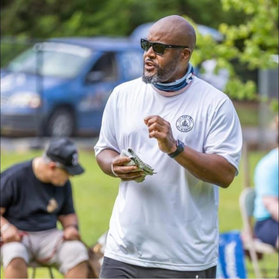 Man in a white t-shirt with a logo, speaking and gesturing during a dog training session, surrounded by individuals in a training environment, emphasizing hands-on learning in dog trainer certification.